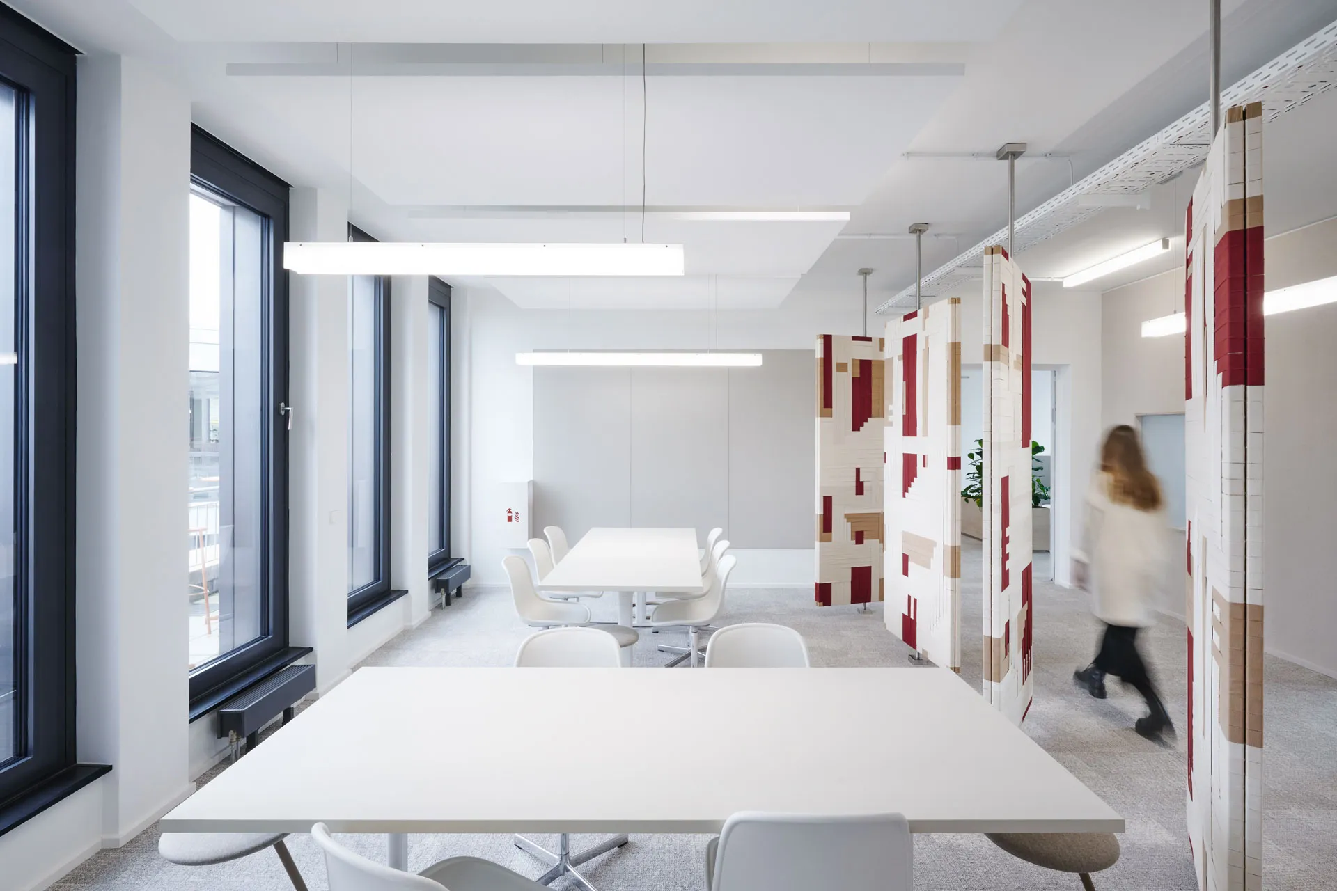 The 'Spinning Jenny,' a project created by Jannis Schäfer Studio in collaboration with INpuls interior studio for Acterience. The image shows a modern office space with sound panels featuring woven graphics made from colored cotton bands in red, white, and beige. The panels are suspended from the ceiling and can spin, adding a dynamic and artistic element to the space.