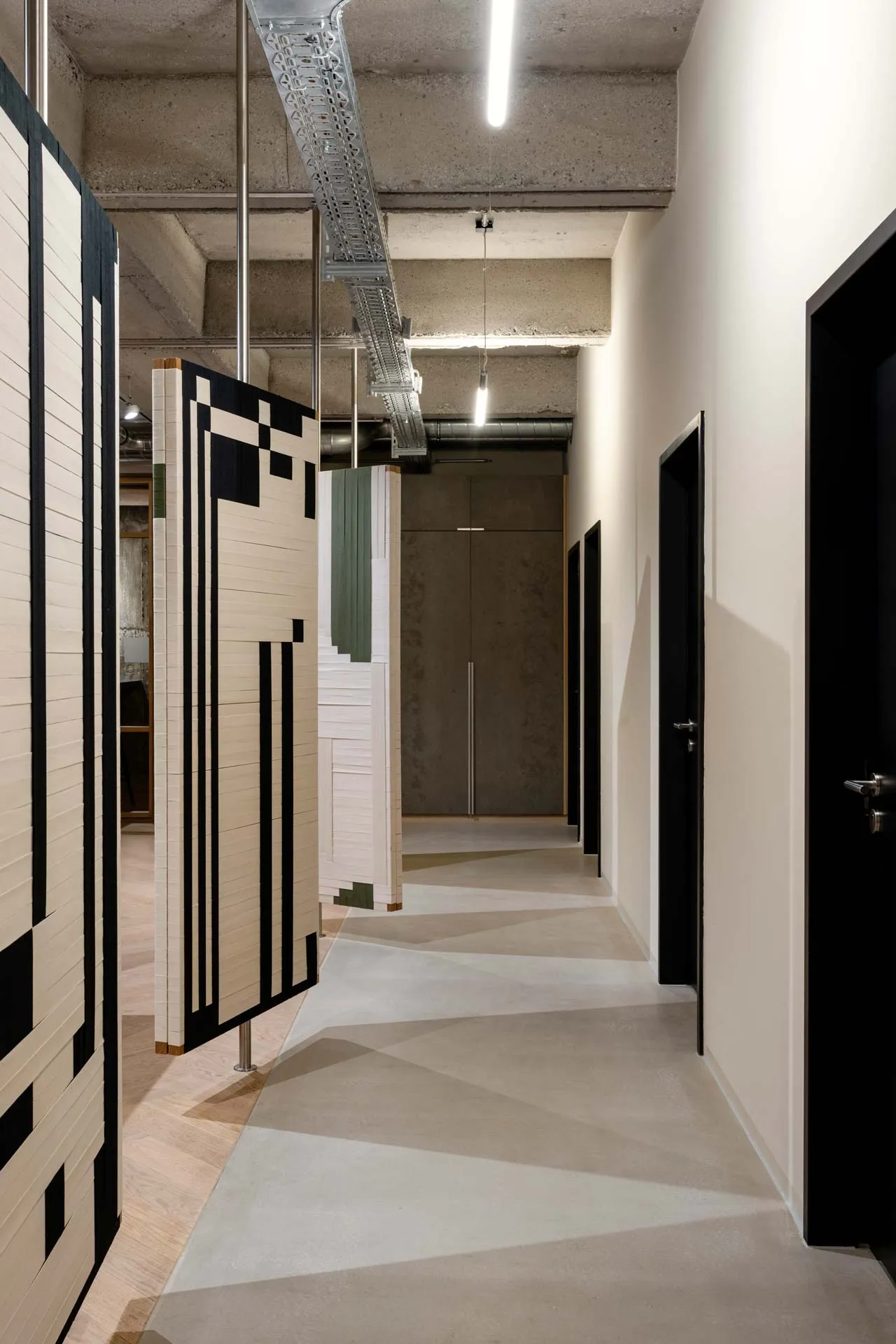 The 'Spinning Jenny,' a project created by Jannis Schäfer Studio in collaboration with INpuls interior studio for KBC Kemény Boehme. The image shows a modern hallway with sound panels featuring woven graphics made from colored cotton bands in black, white, and green. The panels are suspended from the ceiling and can spin, adding a dynamic and artistic element to the space.