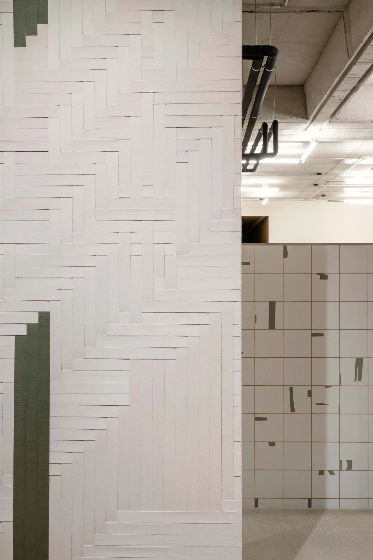 The 'Spinning Jenny,' a project created by Jannis Schäfer Studio in collaboration with INpuls interior studio for KBC Kemény Boehme. The image shows a modern hallway with sound panels featuring woven graphics made from colored cotton bands in black, white, and green. The panels are suspended from the ceiling and can spin, adding a dynamic and artistic element to the space.